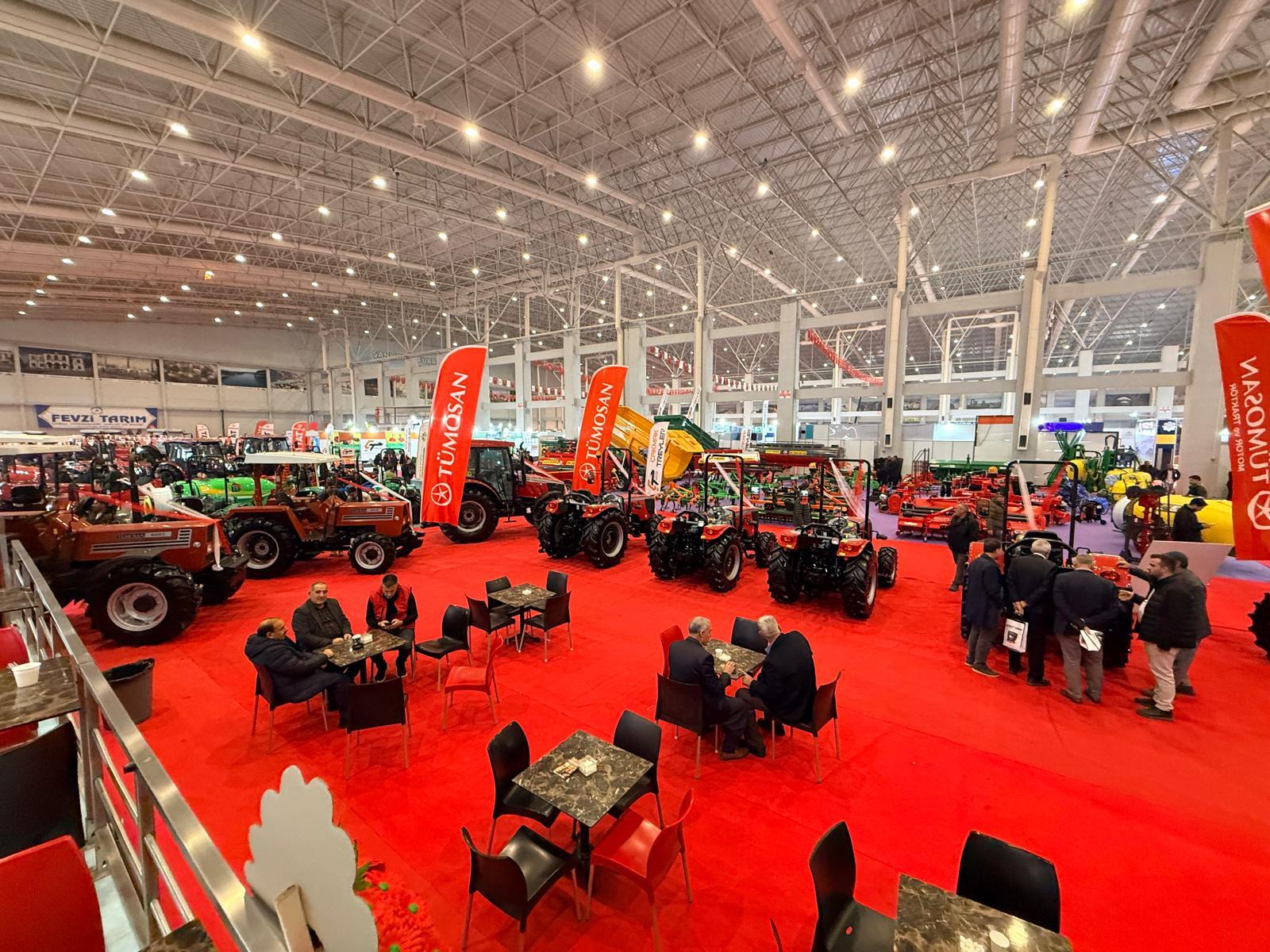 Indoor agricultural fair with tractors on red carpet, people browsing exhibits and sitting at tables.