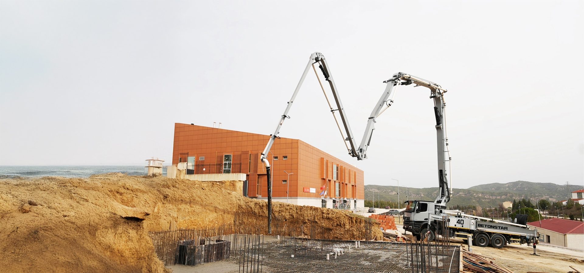 A concrete pump truck pouring concrete into a foundation at a construction site near an orange building and the sea.