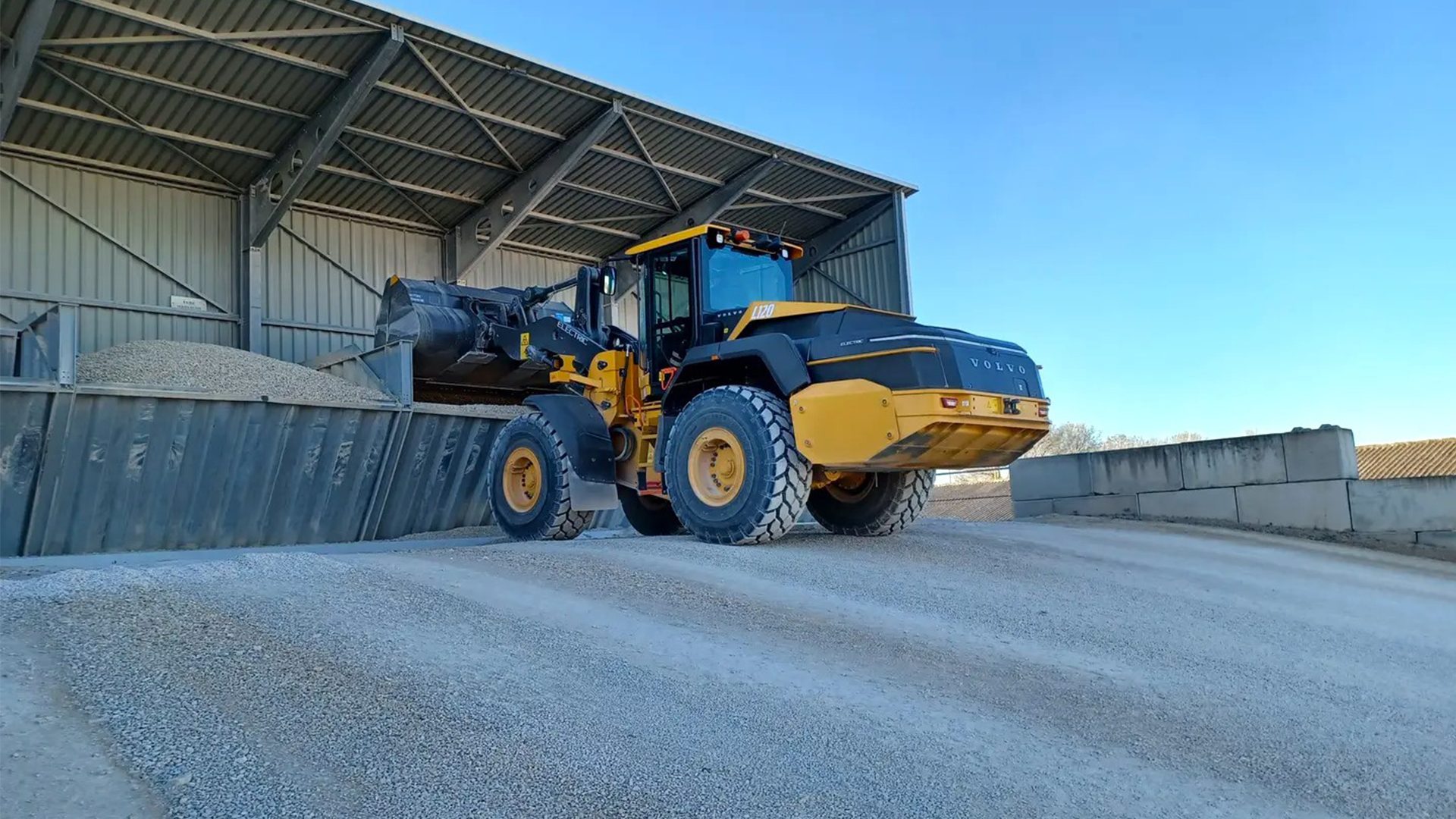 A yellow Volvo wheel loader loads gravel from a pile under a large shed.