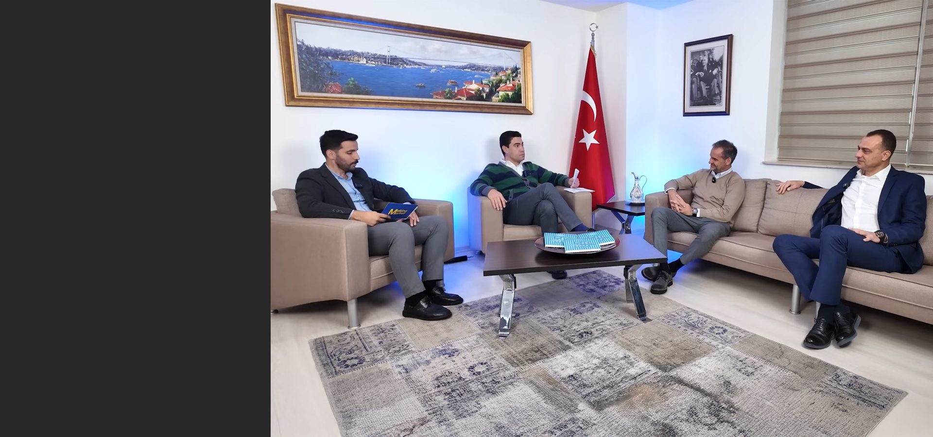 Four men sit in a modern lounge with a Turkish flag and a Bosphorus painting on the wall.