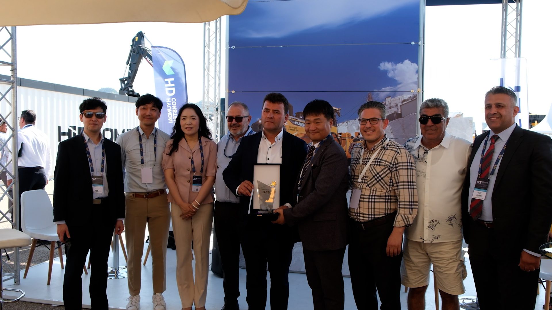 Outdoor group photo of 10 people. Two men in center hold a white award.