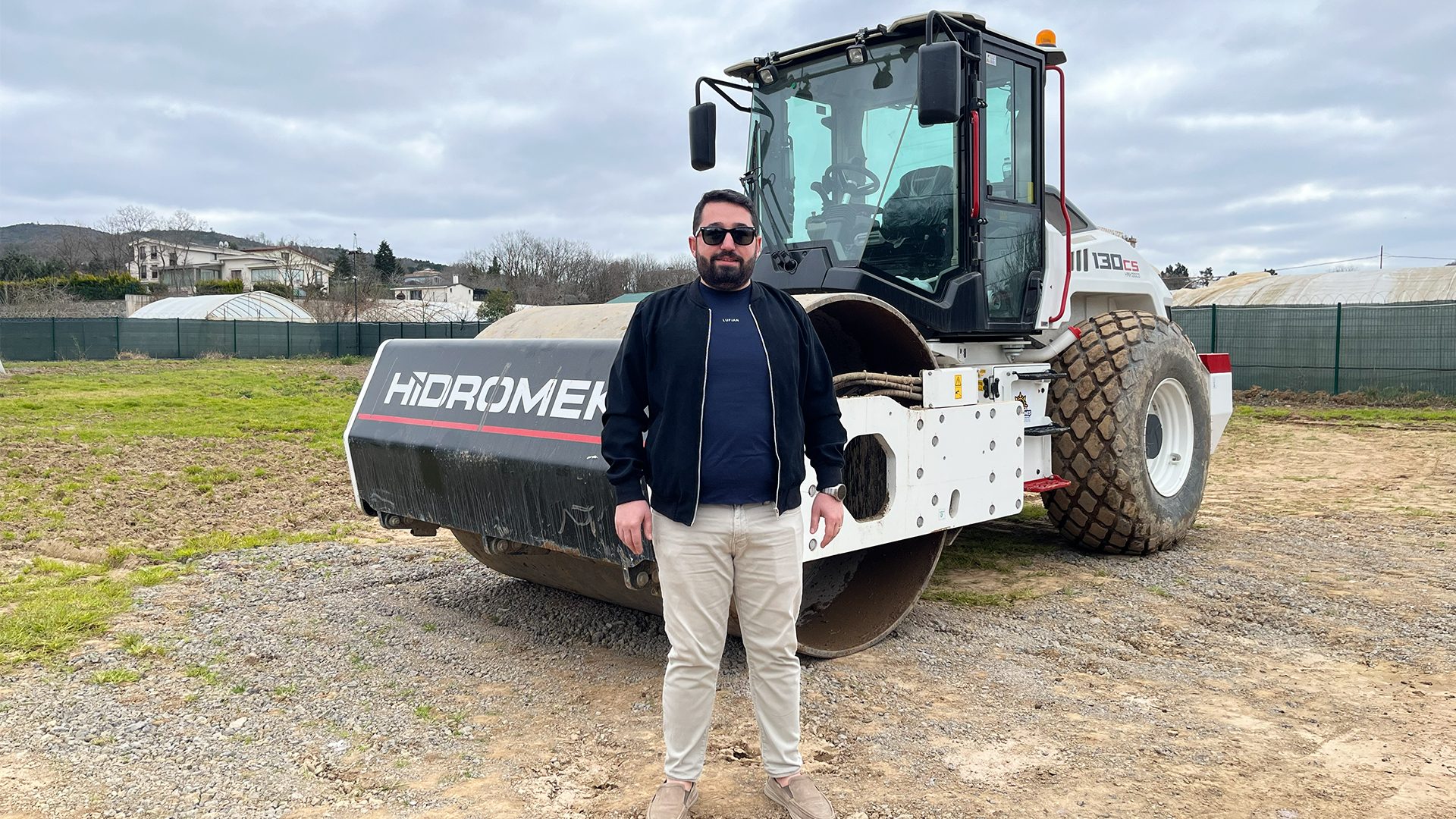Man in sunglasses and jacket standing confidently in front of a large HIDROMEK road roller on a construction site.