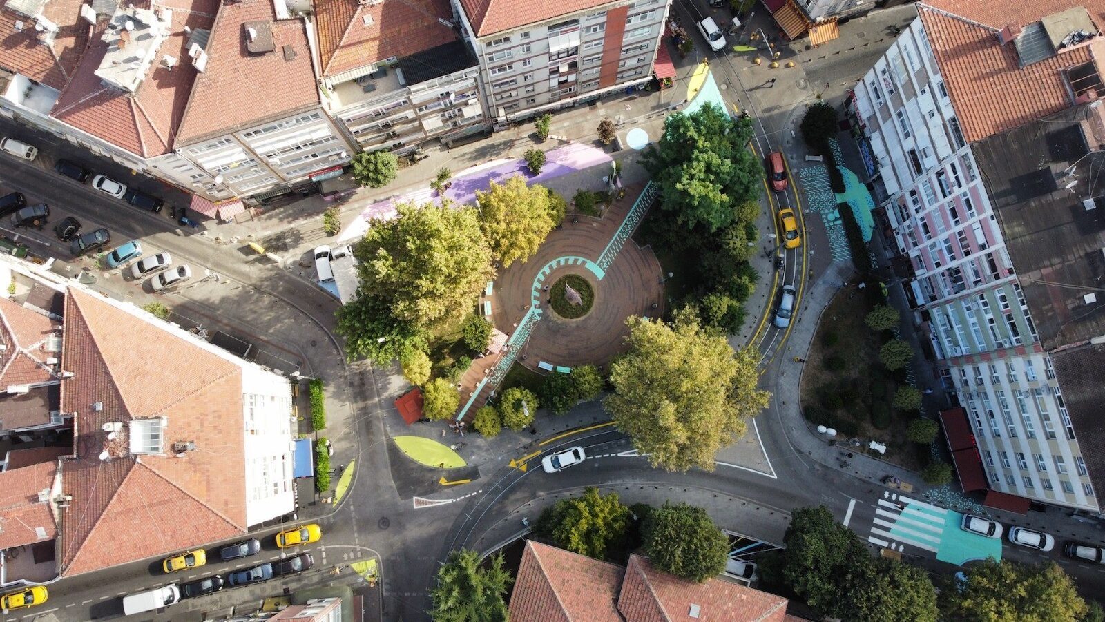 Aerial view: busy city square with a circular park, vibrant street art, buildings, cars, and trees.