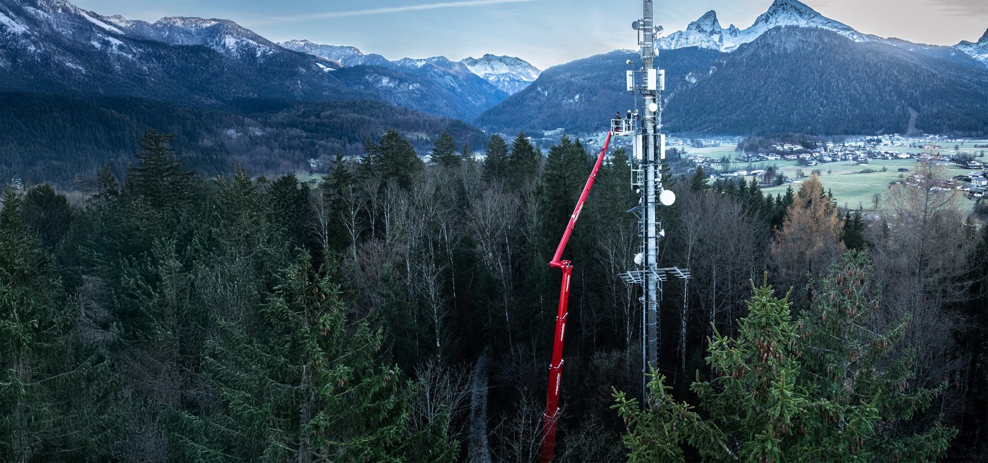 Workers on a red boom lift maintaining a cell tower in a forested mountain landscape with a village.