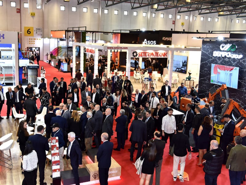 Attendees networking at a busy trade show with various exhibition booths and industrial machinery.