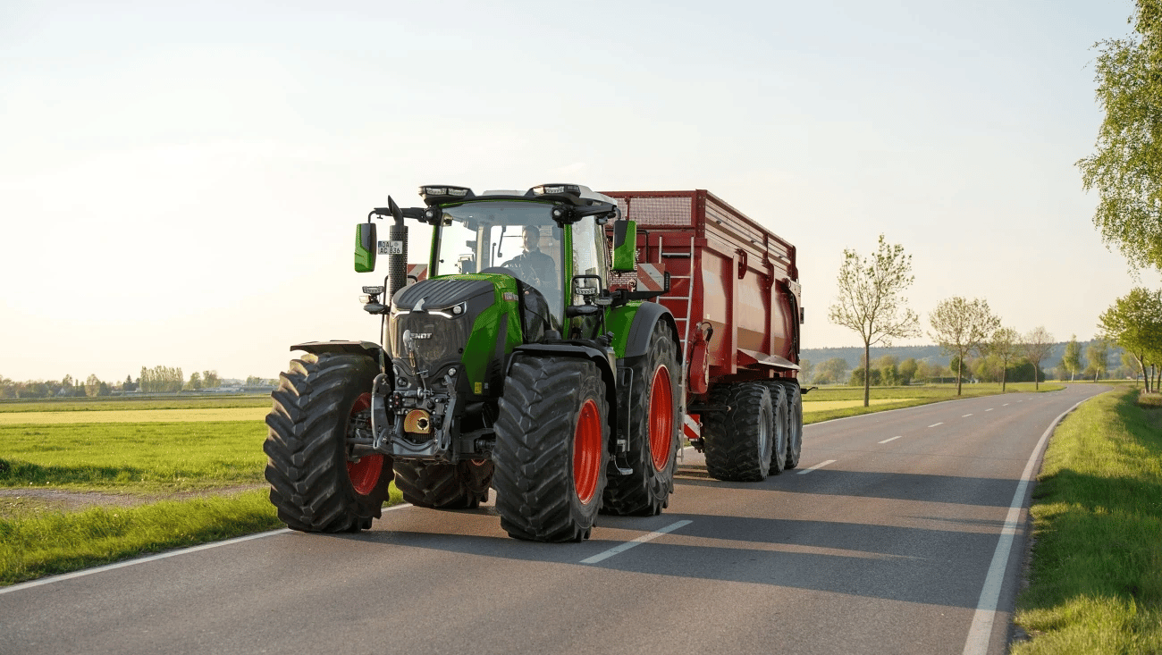 Green Fendt tractor pulling a red trailer on a rural road with fields and trees.