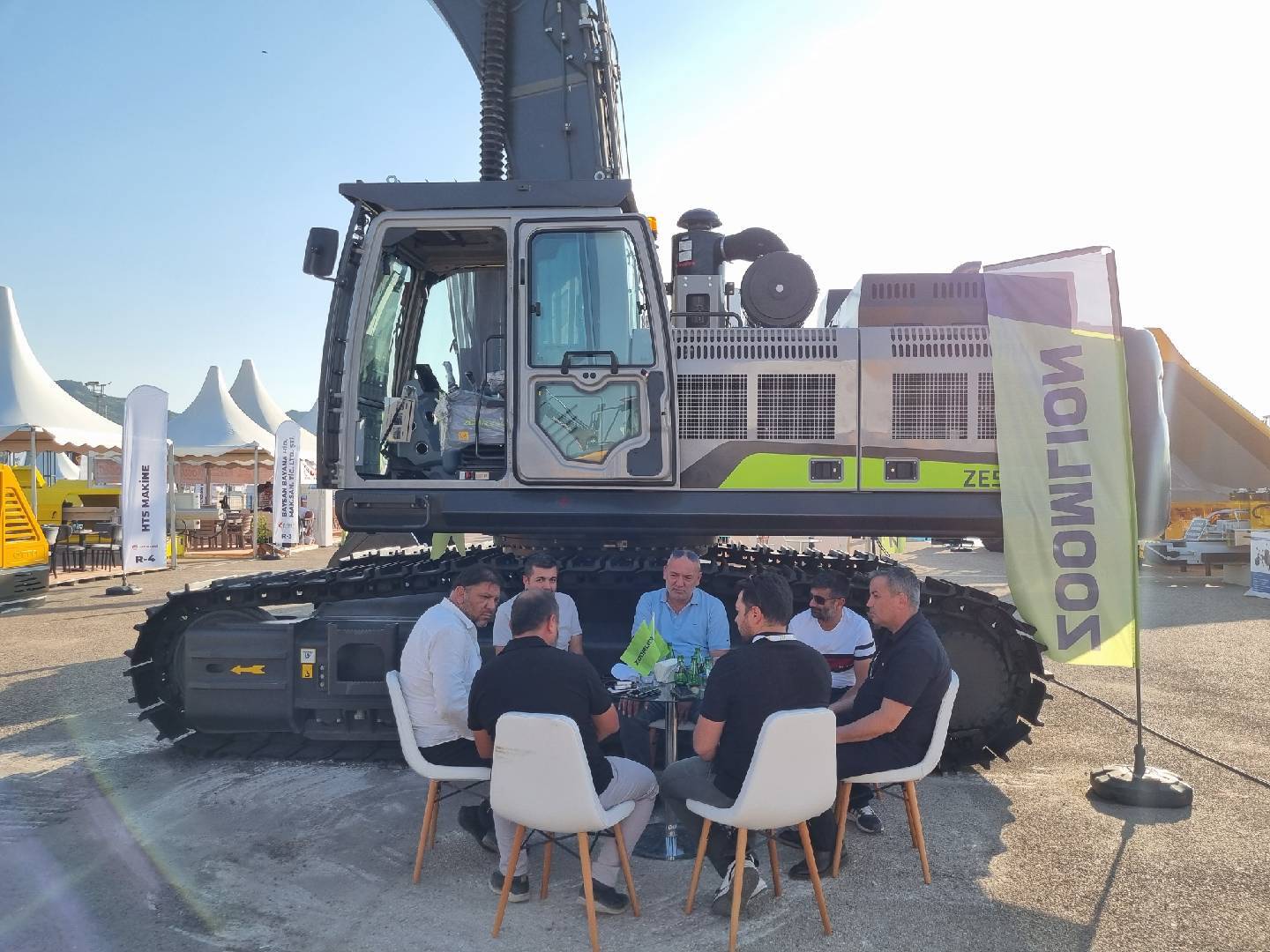 Six men meeting next to a ZOOMLION excavator at an outdoor trade fair.