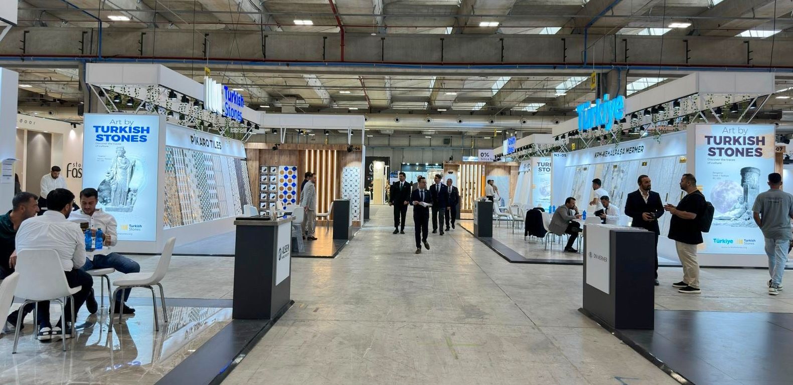 Indoor trade show with "Turkish Stones" and "Pikaro Tiles" exhibits and attendees.