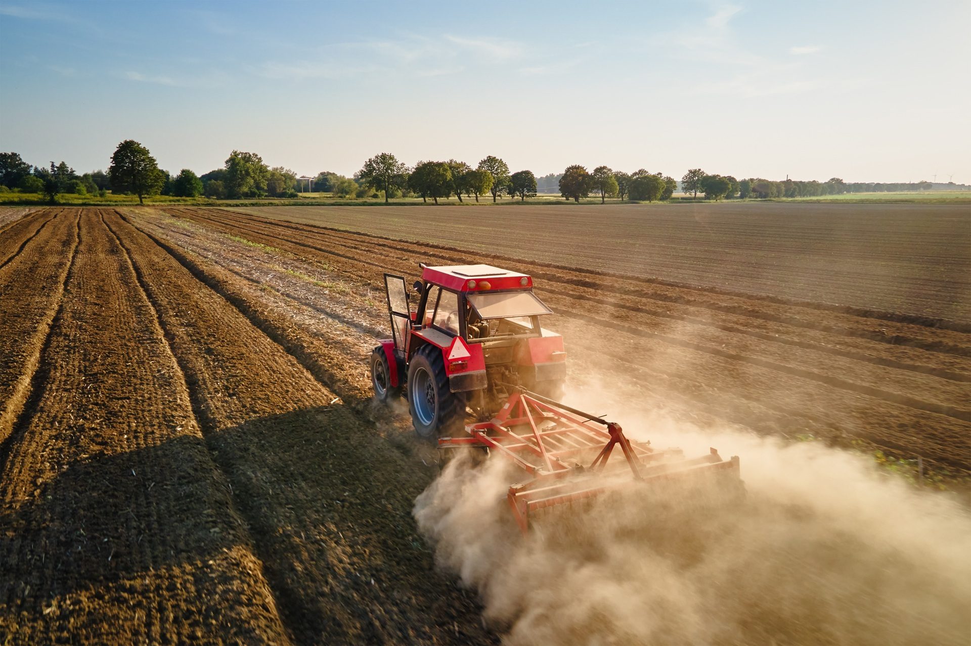A red tractor plows a field, kicking up a large cloud of dust under a clear sky.
