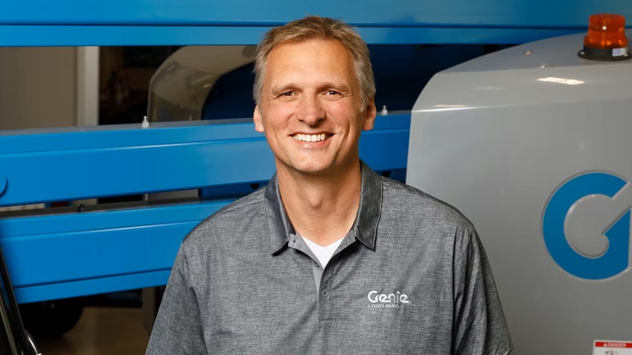Smiling man in a grey Genie polo shirt standing in front of blue and grey industrial machinery.