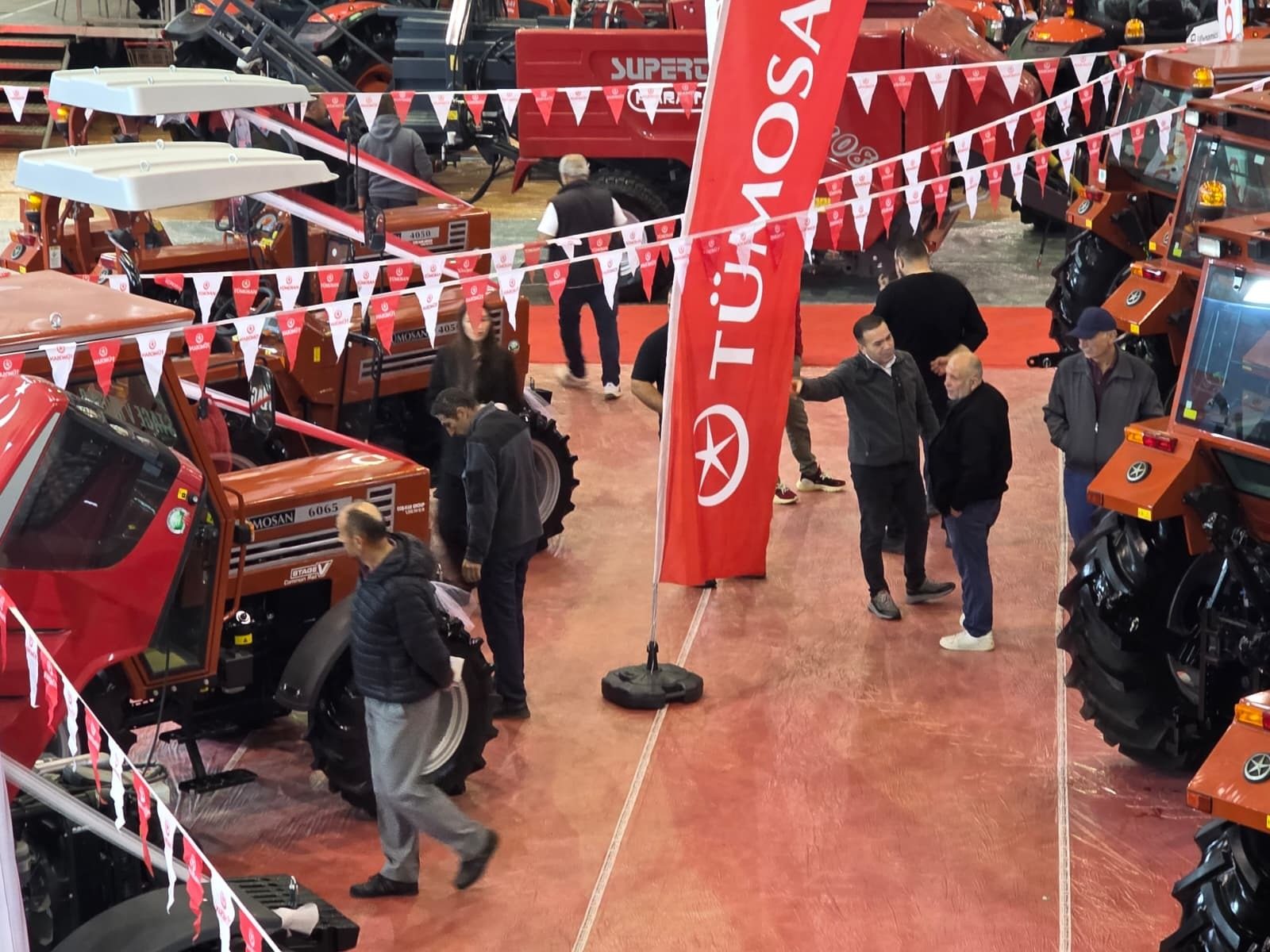 Attendees view tractors at an indoor exhibition with a red TÜMOSAN banner and decorative bunting.