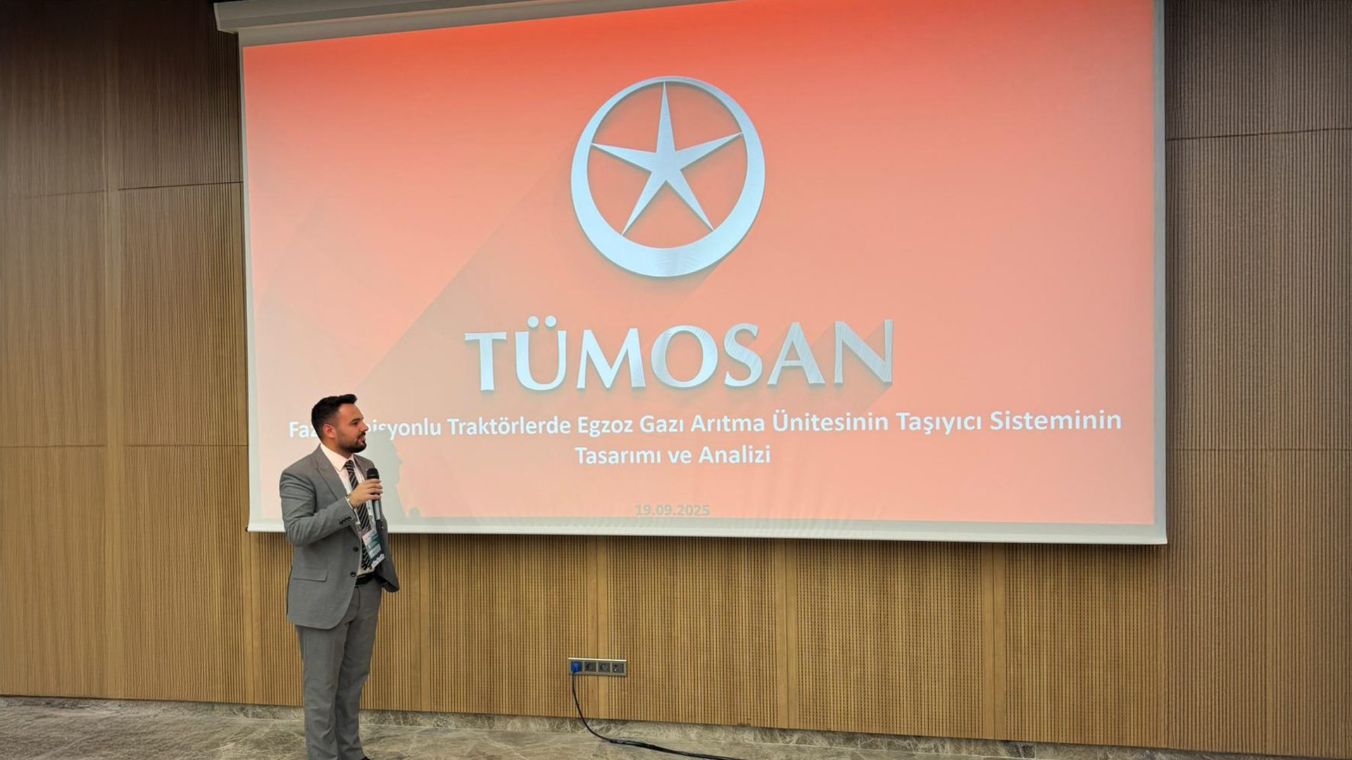 Man presenting at a TÜMOSAN event, screen shows logo and Turkish text on exhaust gas treatment system.