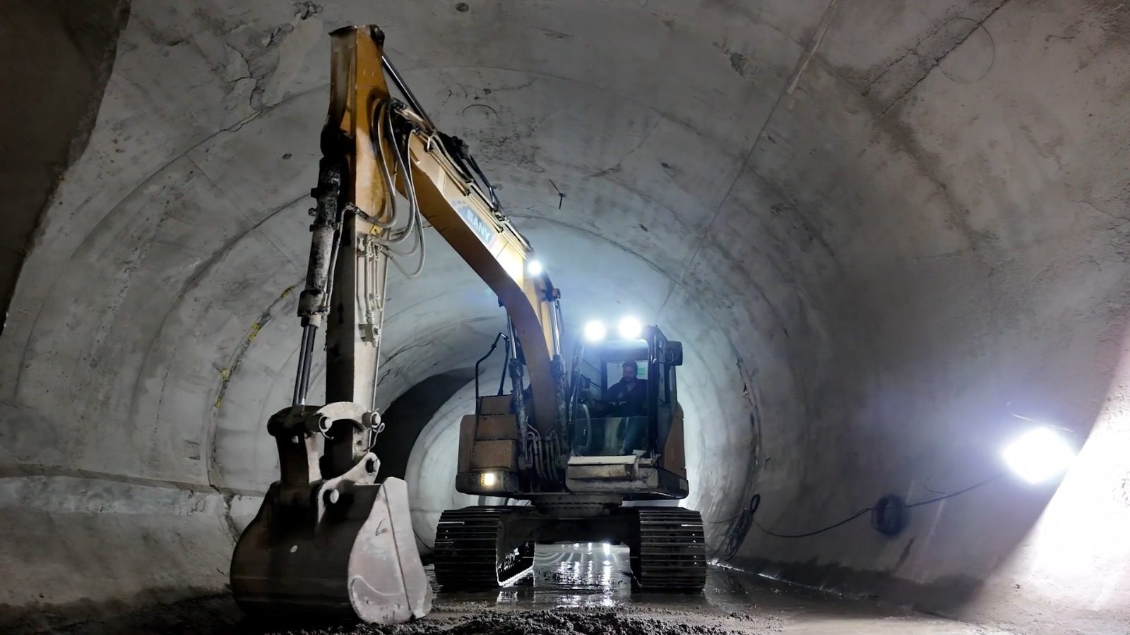 An excavator with an operator inside a concrete tunnel, illuminated by bright lights.