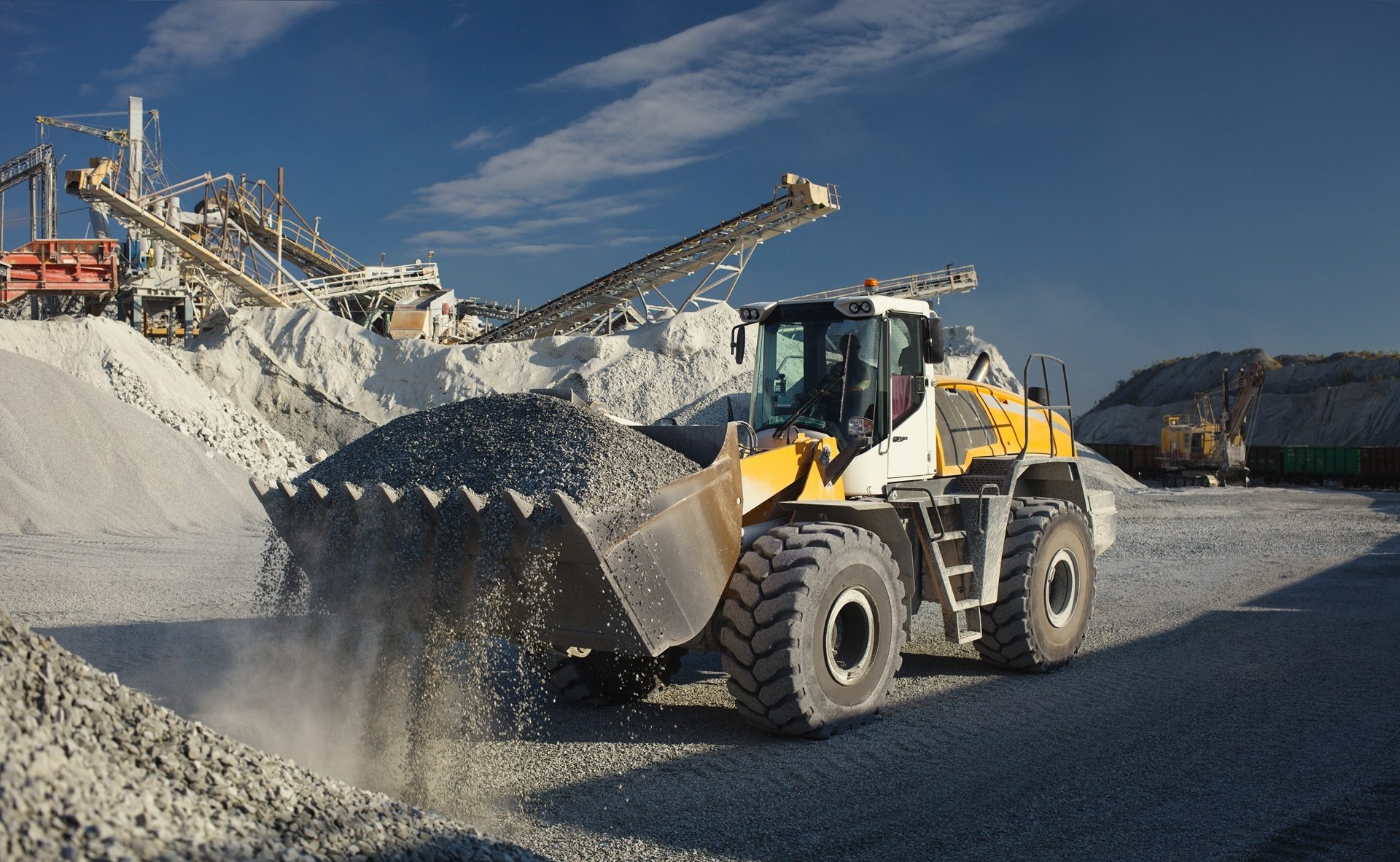 A yellow front-end loader scooping gravel at a quarry with crushing machinery under a blue sky.