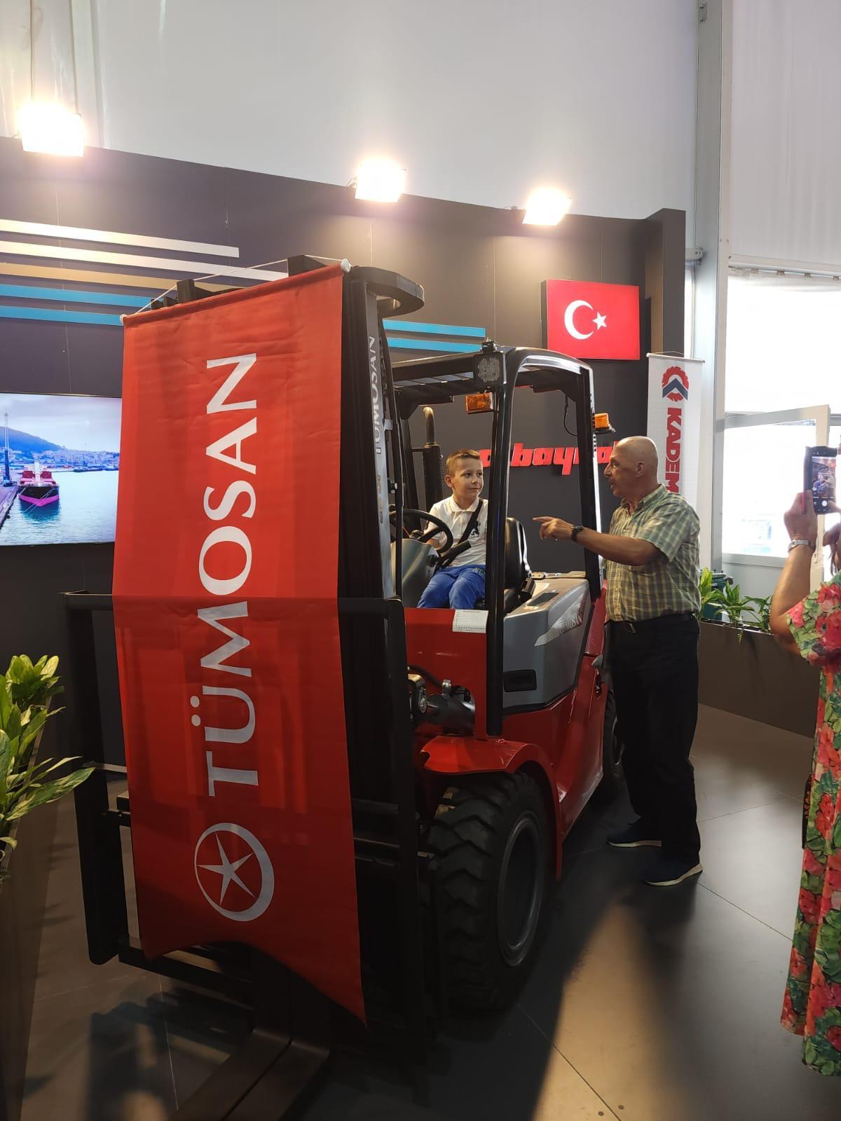 Boy in a red TÜMOSAN forklift, man pointing, Turkish flag in background.