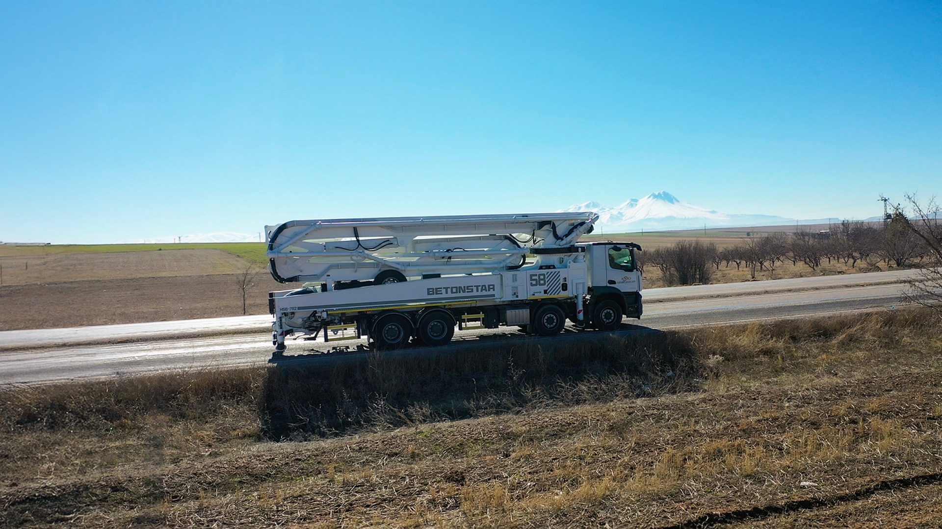 White Betonstar concrete pump truck on a road with fields and snow-capped mountains in the background.
