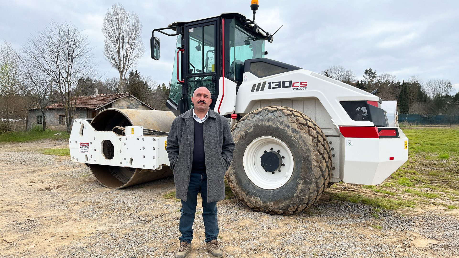 A man with a mustache in a gray coat stands in front of a white road roller on a dirt path.