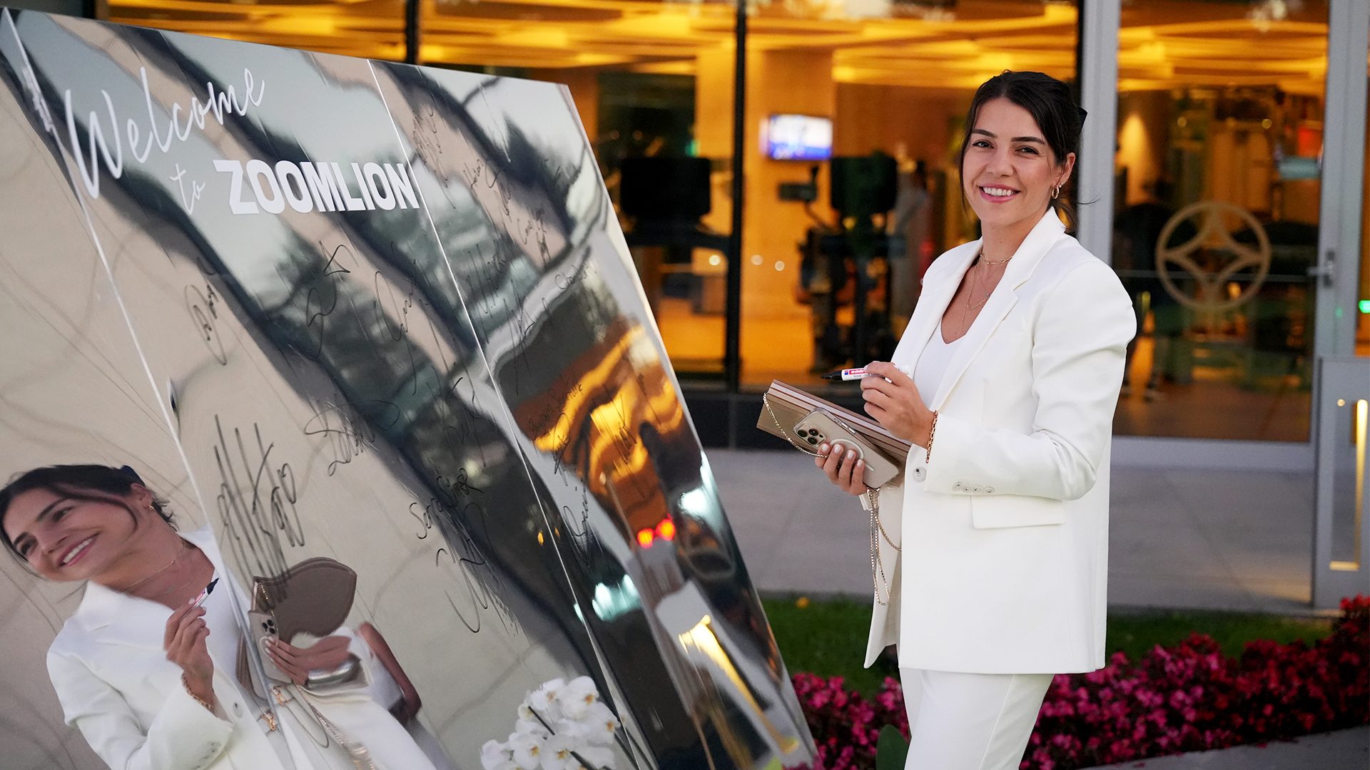 Smiling woman in white by a mirrored "Welcome to ZOOMLION" sign covered in signatures.
