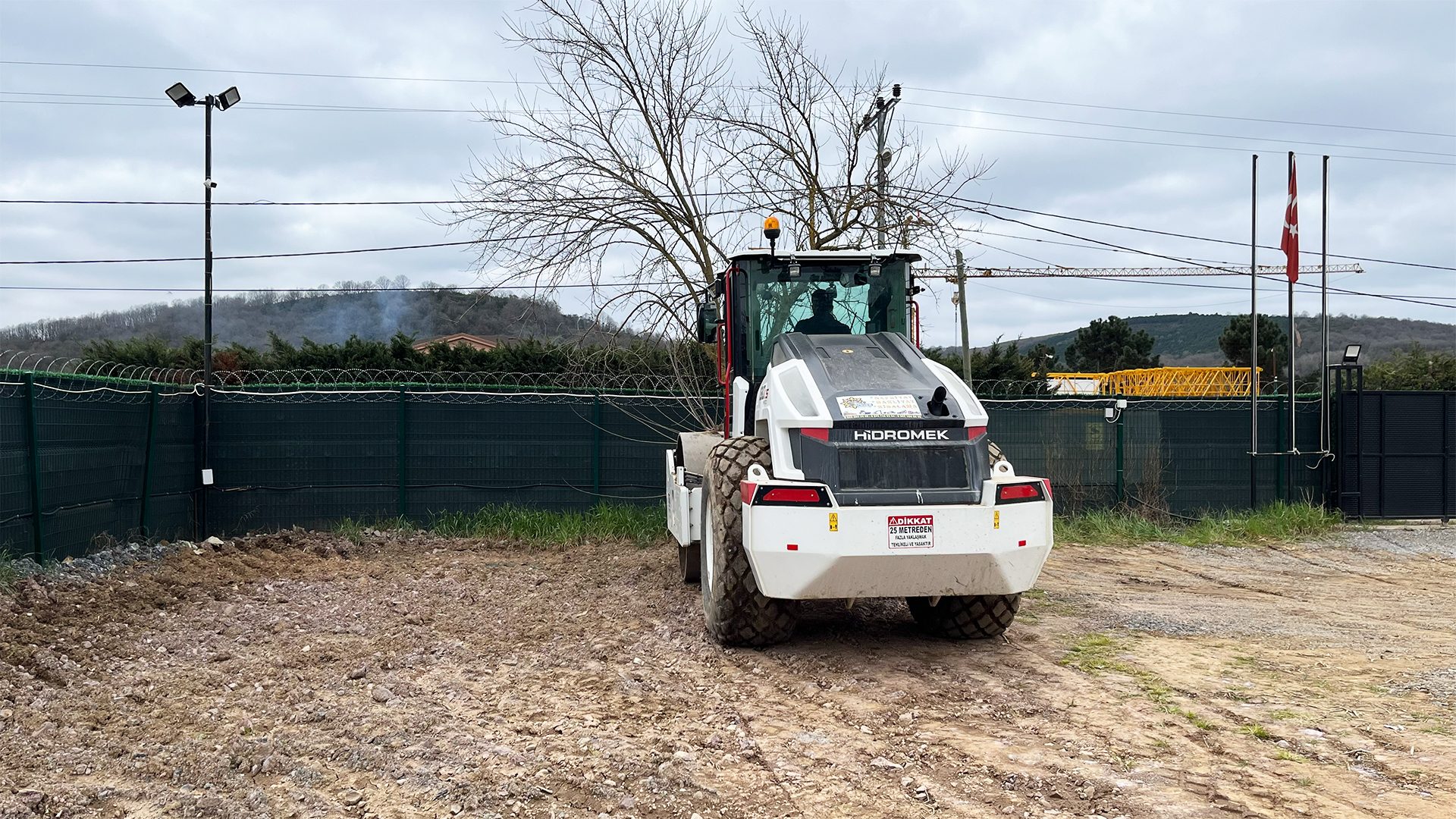 A white and black Hidromek roller parked on a dirt construction site with a fence and bare trees.
