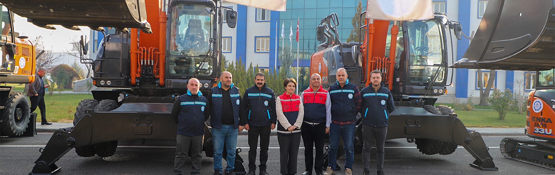 Seven people in vests stand in front of large excavators with a building behind them.