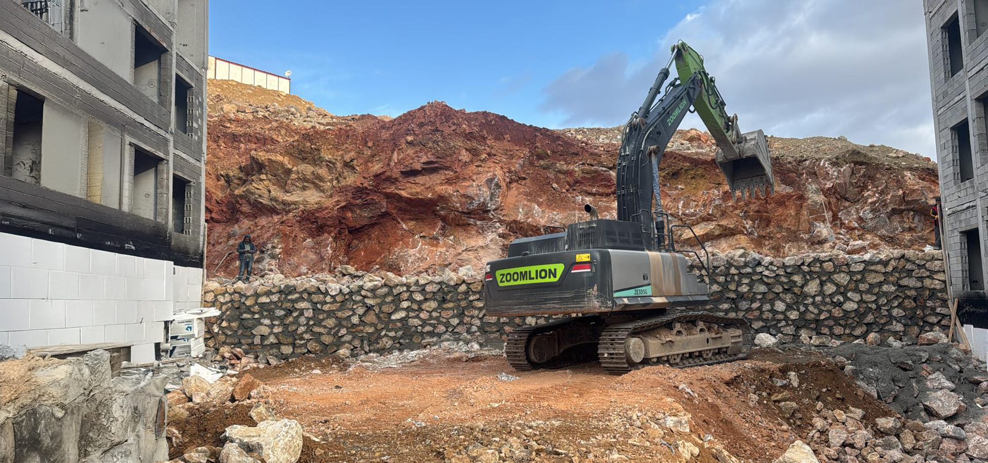 A Zoomlion excavator at a construction site, digging into a red rocky hillside next to unfinished buildings.