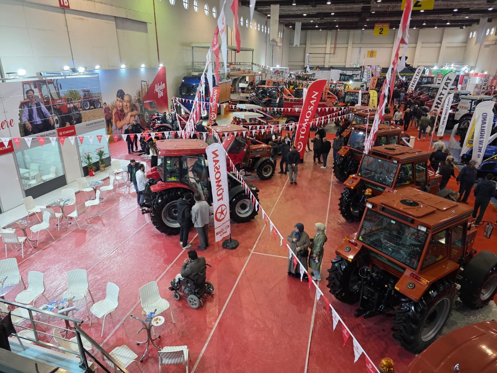Overhead view of a bustling indoor tractor exhibition with red and orange tractors, people, and brand banners.