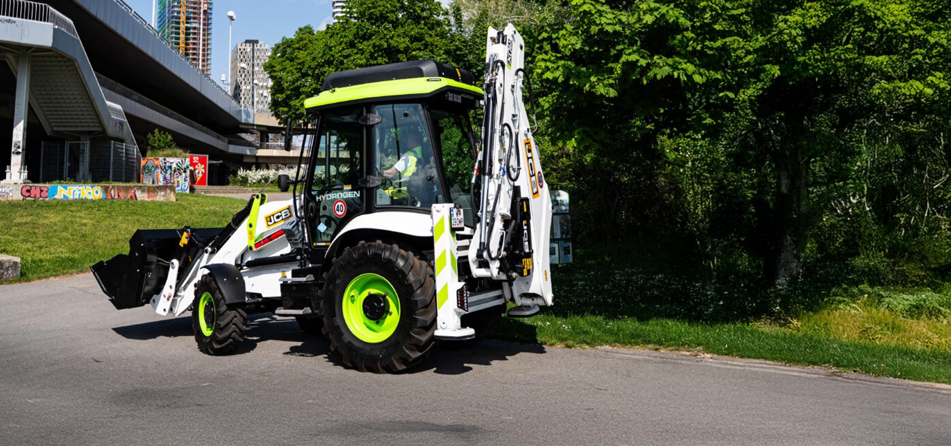 A white and lime green JCB hydrogen-powered backhoe loader on a paved path near a bridge and trees.