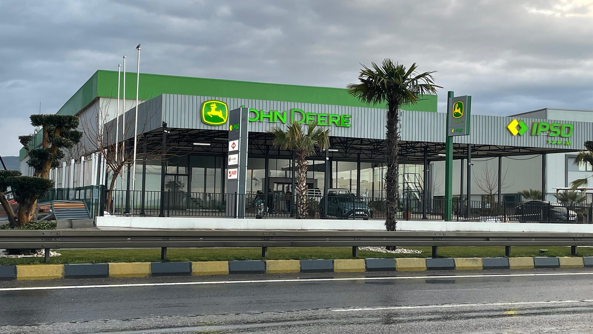 John Deere dealership building with a green roof, palm trees, and a wet road on a cloudy day.