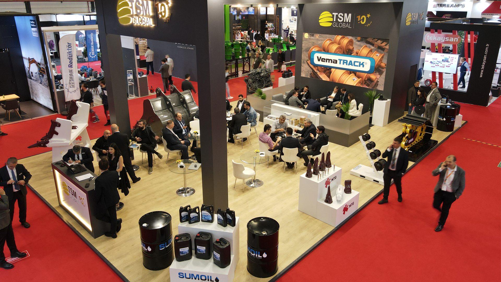 Busy TSM Global booth at a trade show with various product displays, seating areas, and many attendees.