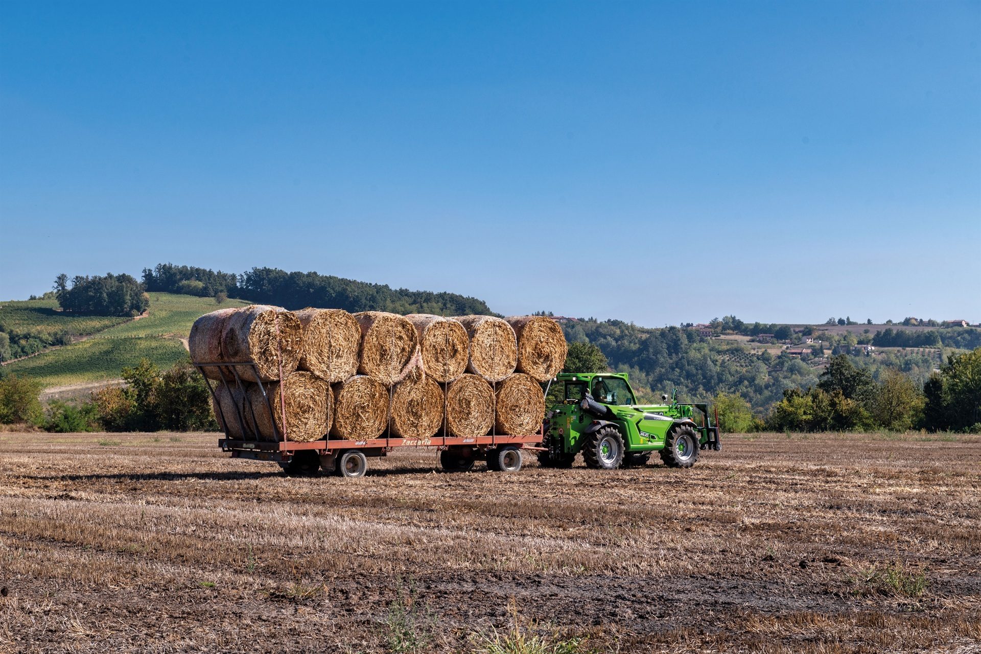 Green telehandler pulls a trailer of hay bales across a dry field under a blue sky.