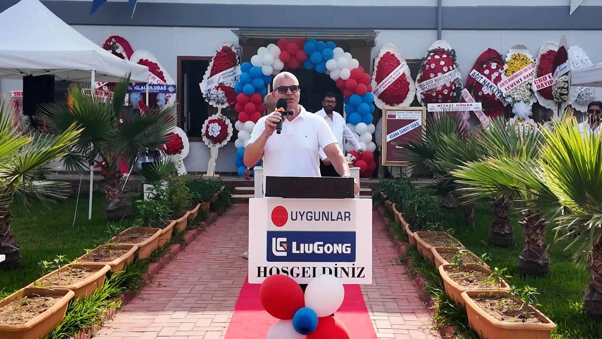 Man speaks at podium for Uygunlar & LiuGong outdoor event, with balloons and floral wreaths.
