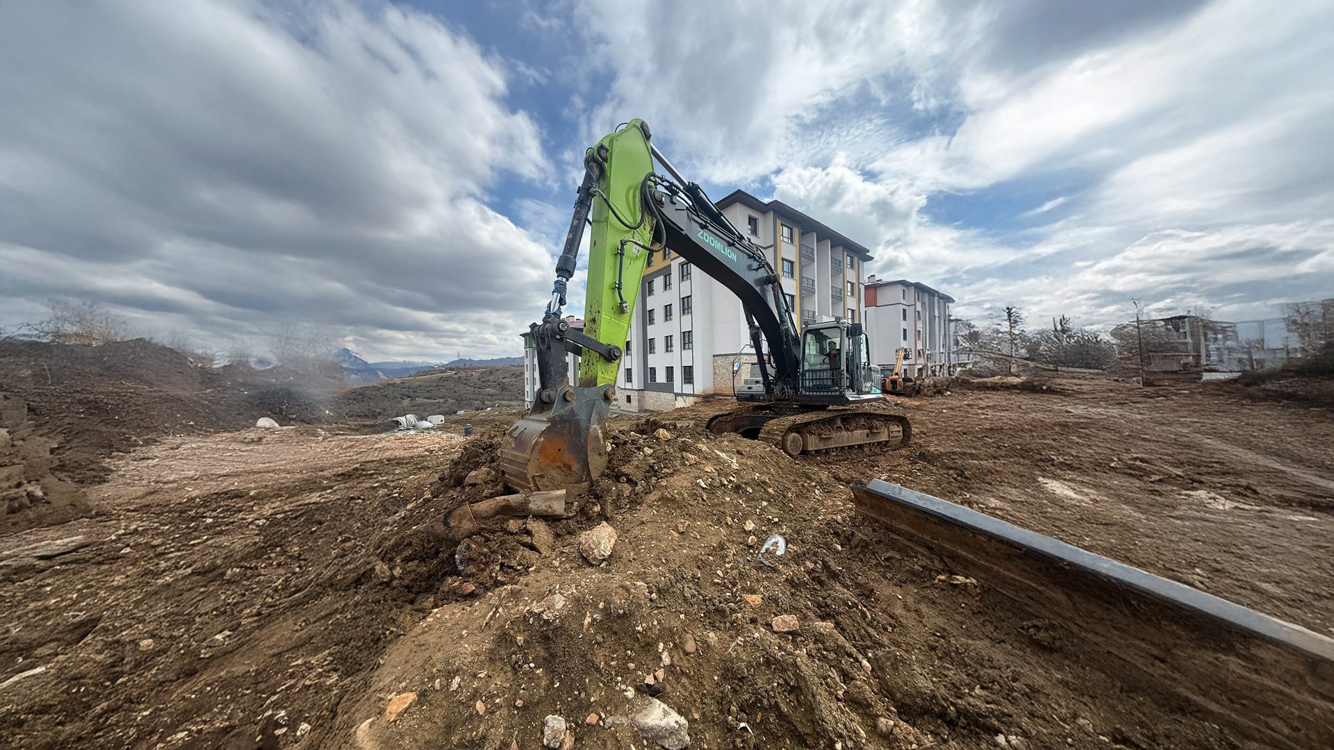 A green and black Zoomlion excavator working on a muddy construction site near apartment buildings under cloudy skies.