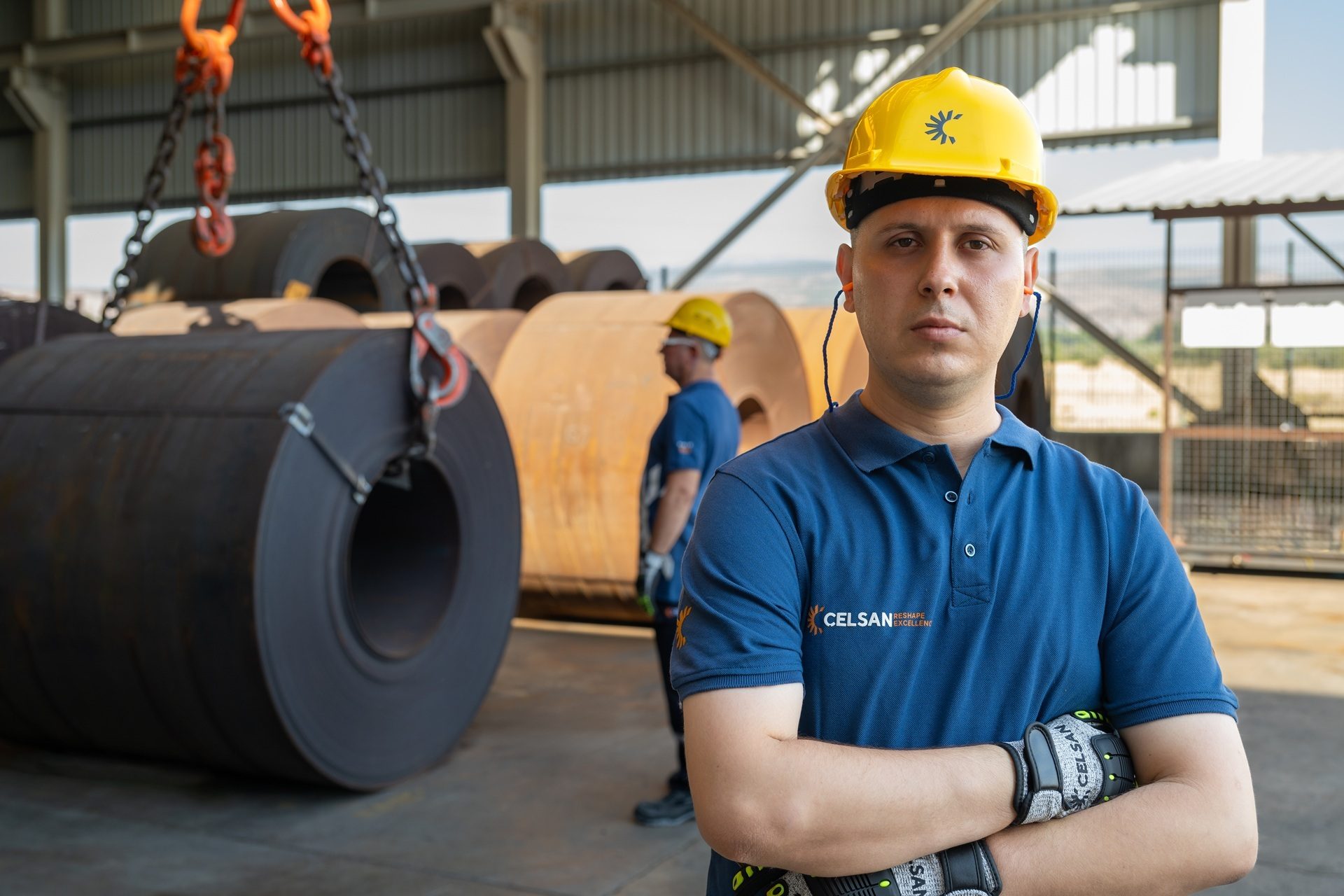 Male CELSAN worker in hard hat and safety gear, arms crossed, in a steel coil warehouse.