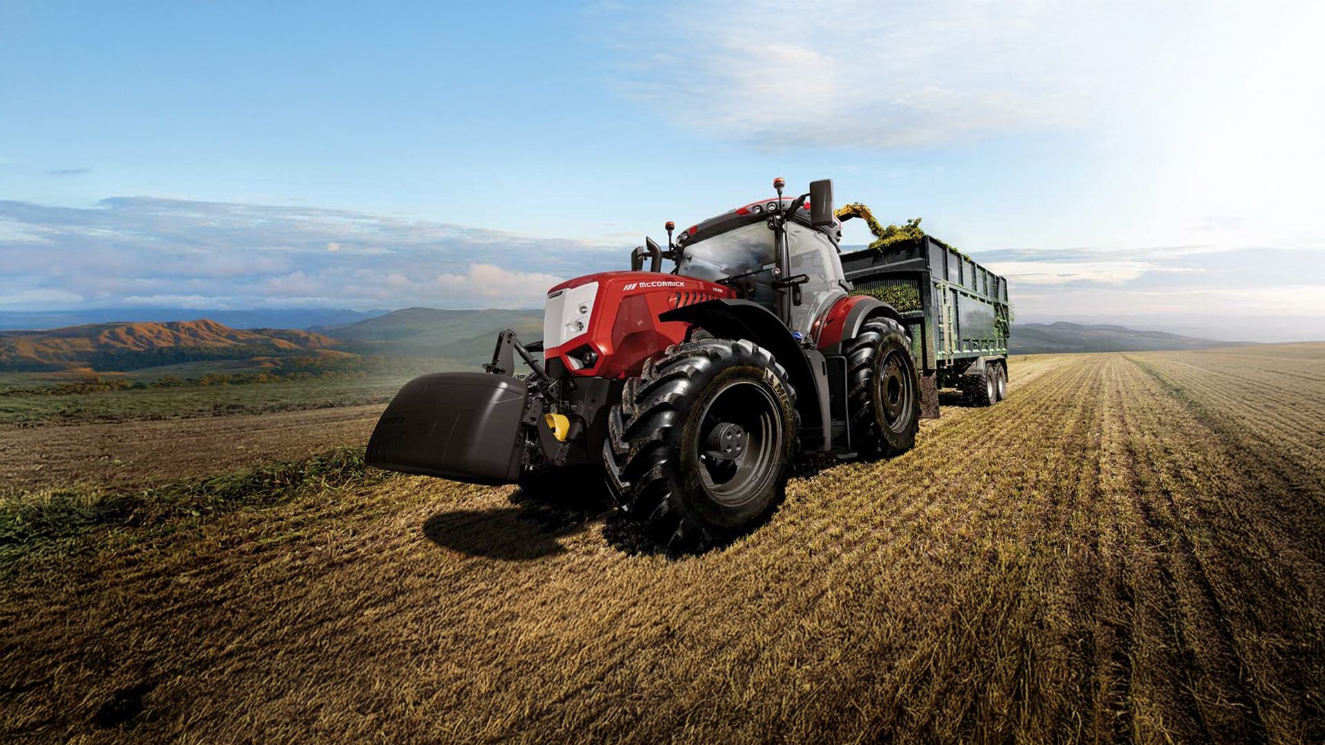 Red McCormick tractor with trailer harvesting crops in a field with distant mountains.