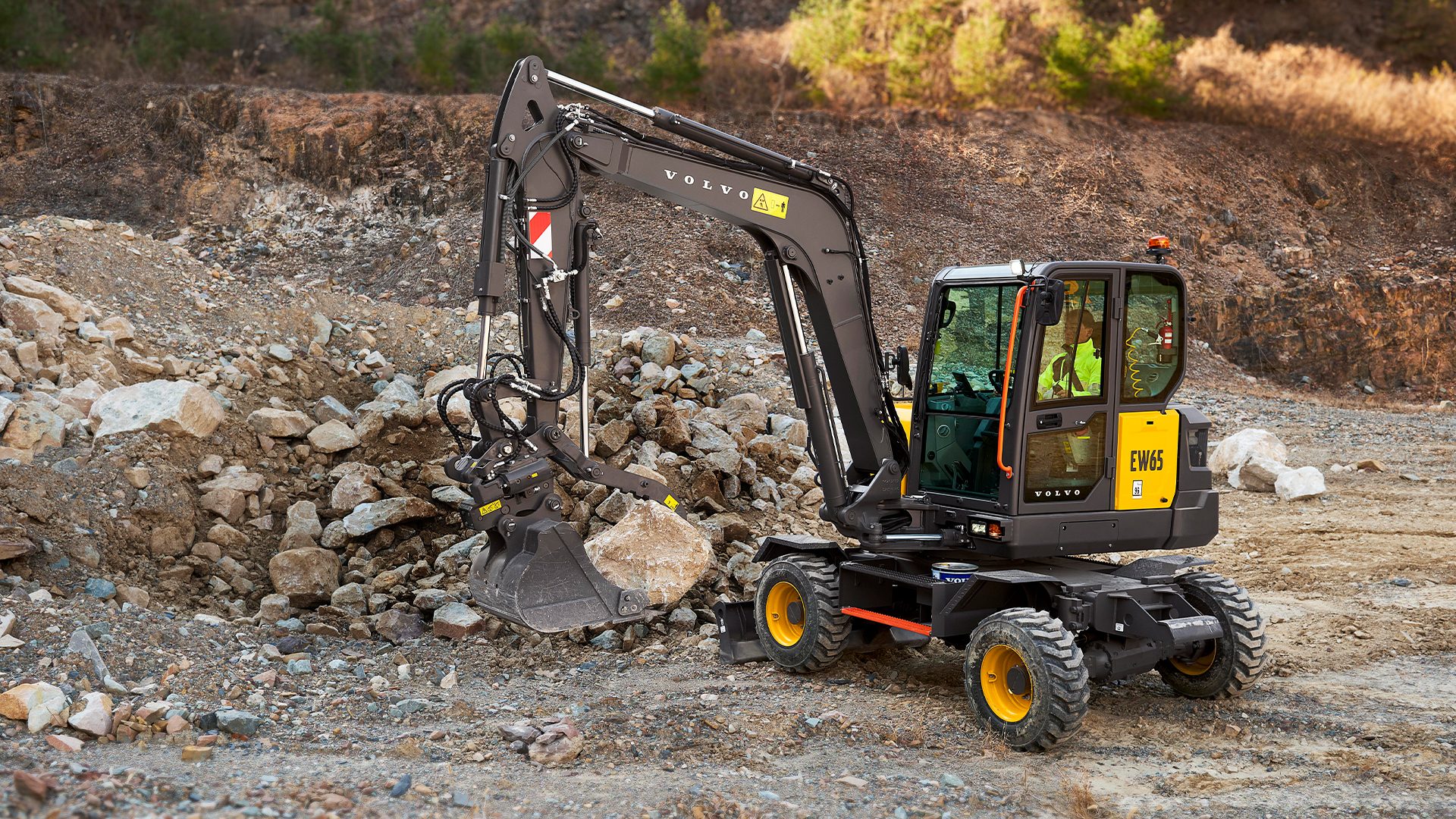 A yellow and grey Volvo EW65 wheeled excavator with an operator, digging rocks in a quarry.