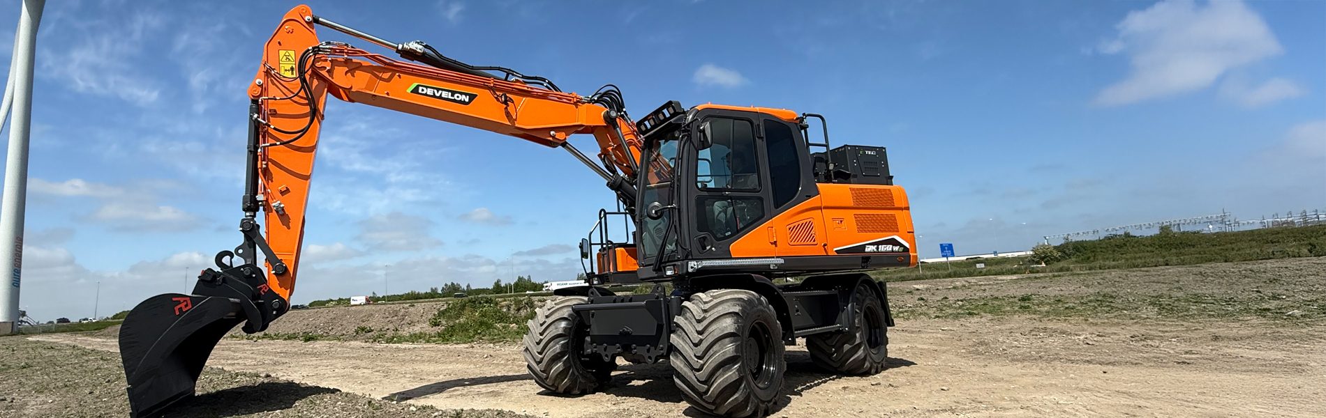 Orange Develon DX 160 W wheeled excavator on dirt ground under a blue sky.