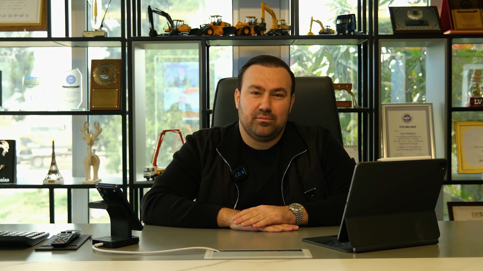 A man in a black jacket sits at a modern desk with a tablet and phone, looking directly at the camera.
