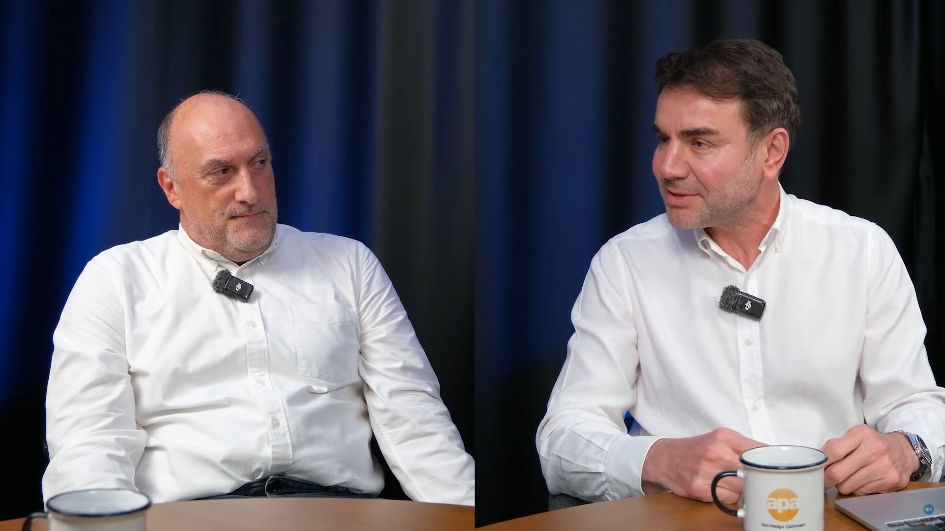 Two men in white shirts, wearing mics, sitting at a table with a dark blue backdrop.
