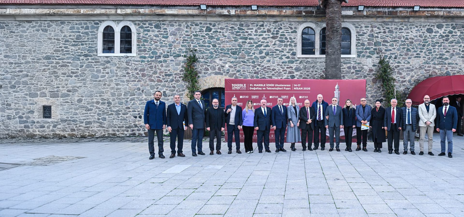 A group of professionals stands in front of a stone building with a banner for the 31st Marble Izmir Fair.