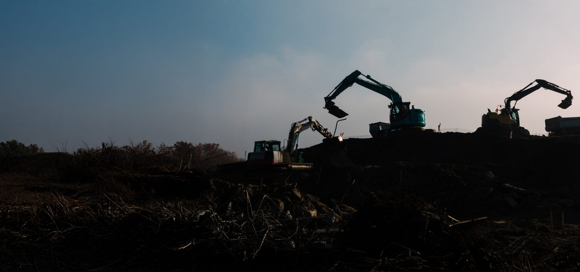 Dark silhouettes of excavators and a dump truck on a construction site under a hazy sky.