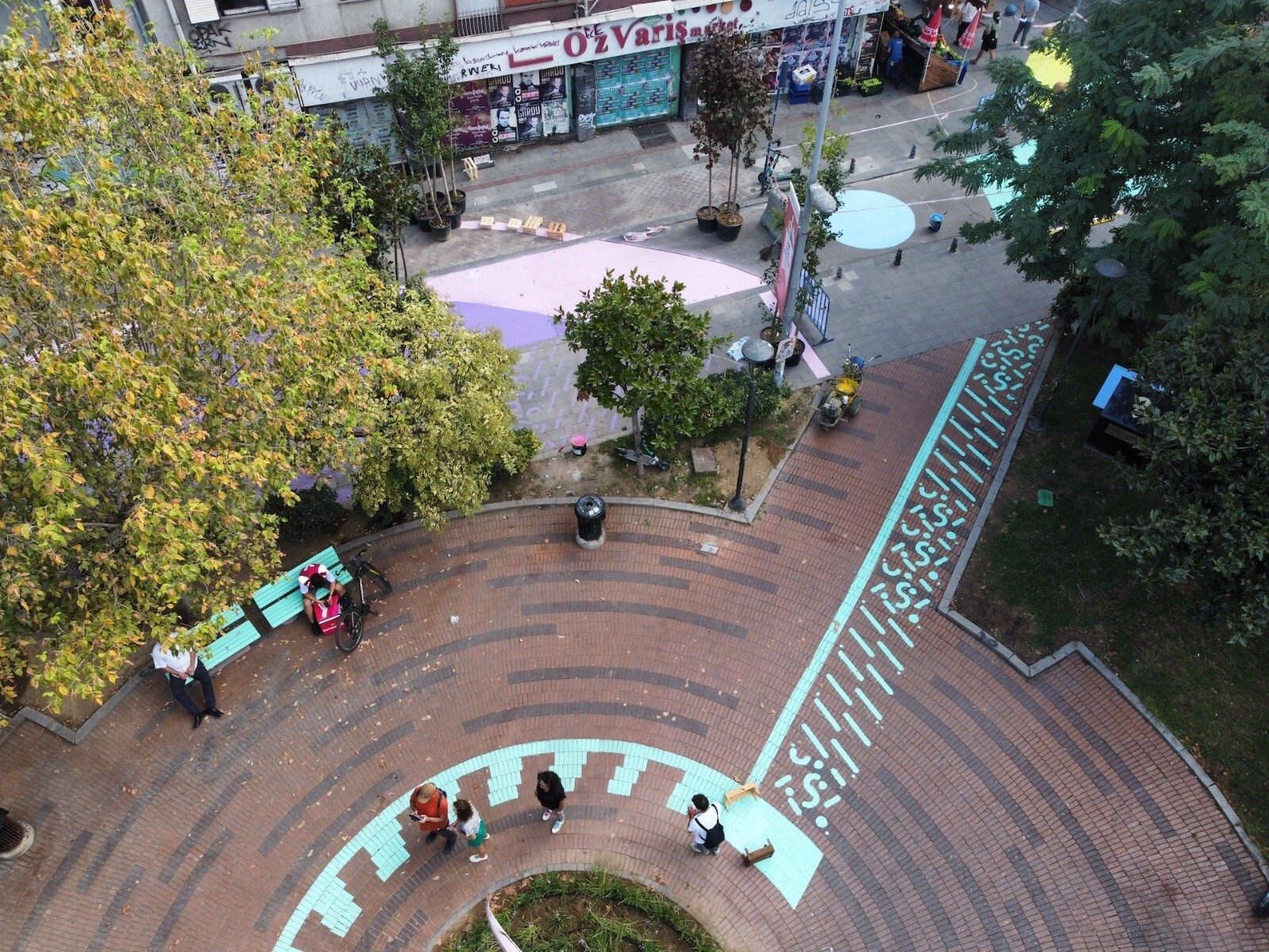 High-angle view of a colorful urban square with people, trees, shops, and geometric patterns on brick paths.