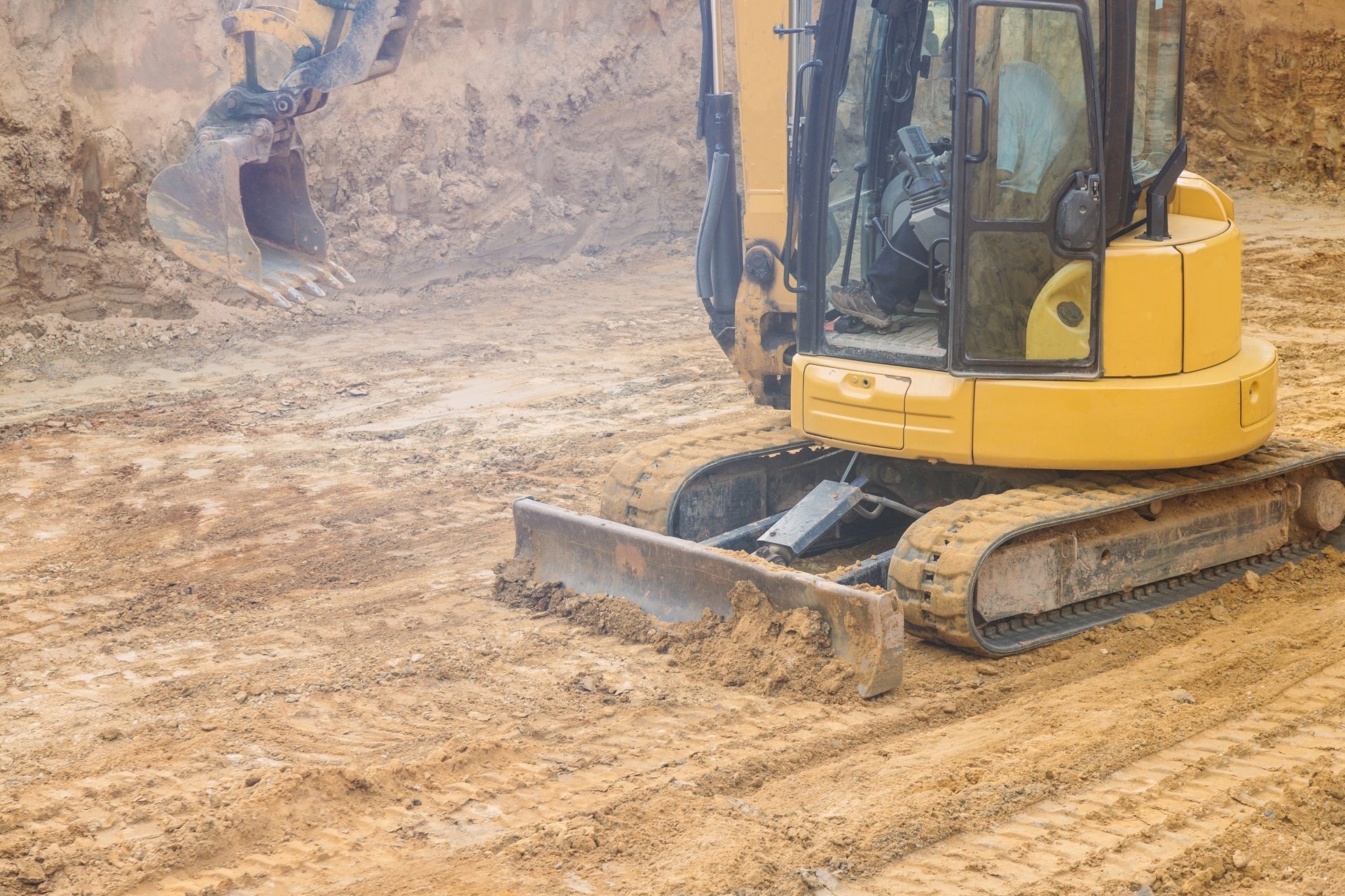 Yellow excavator with raised bucket and blade on sandy construction site.