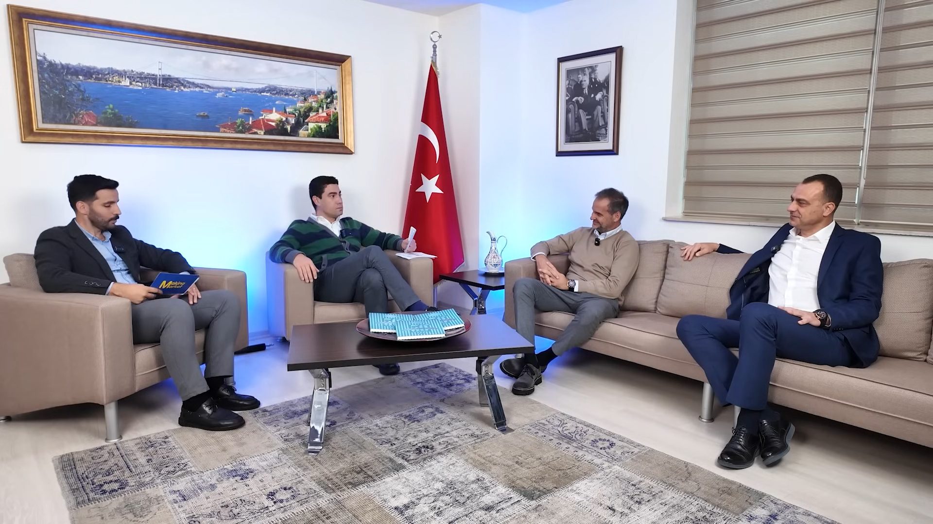 Four men discuss in a studio with a Turkish flag and city painting.