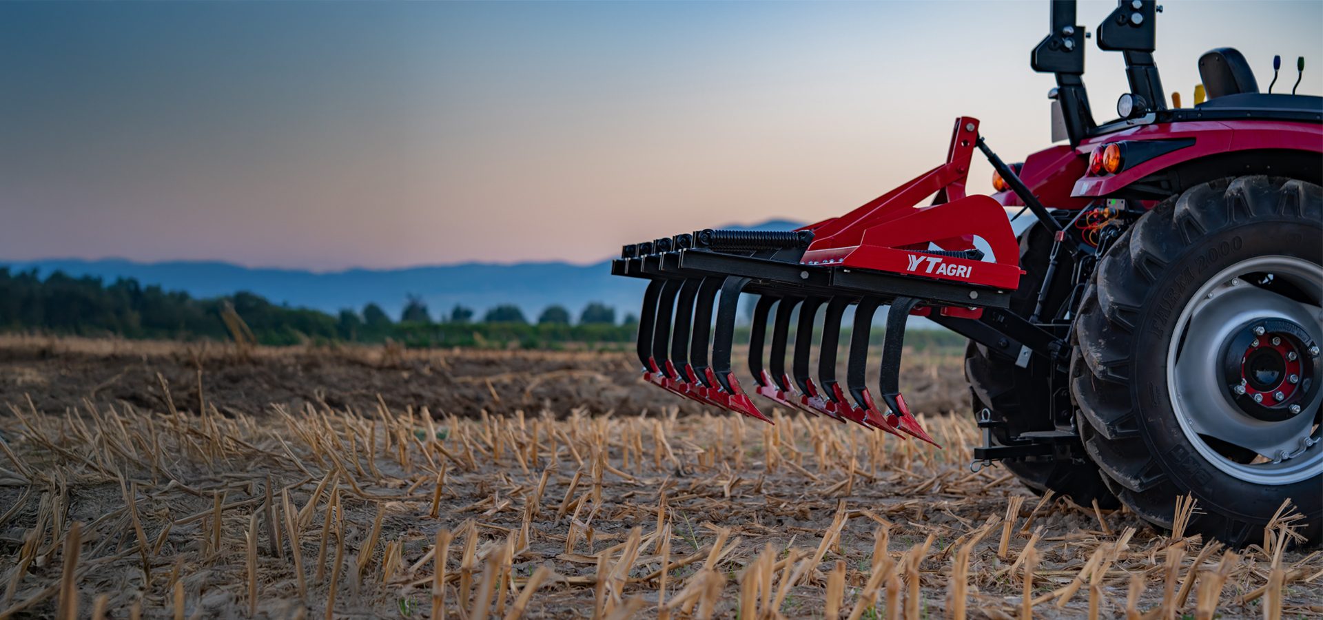 Red agricultural implement attached to a tractor in a stubble field at dusk, with distant hills.