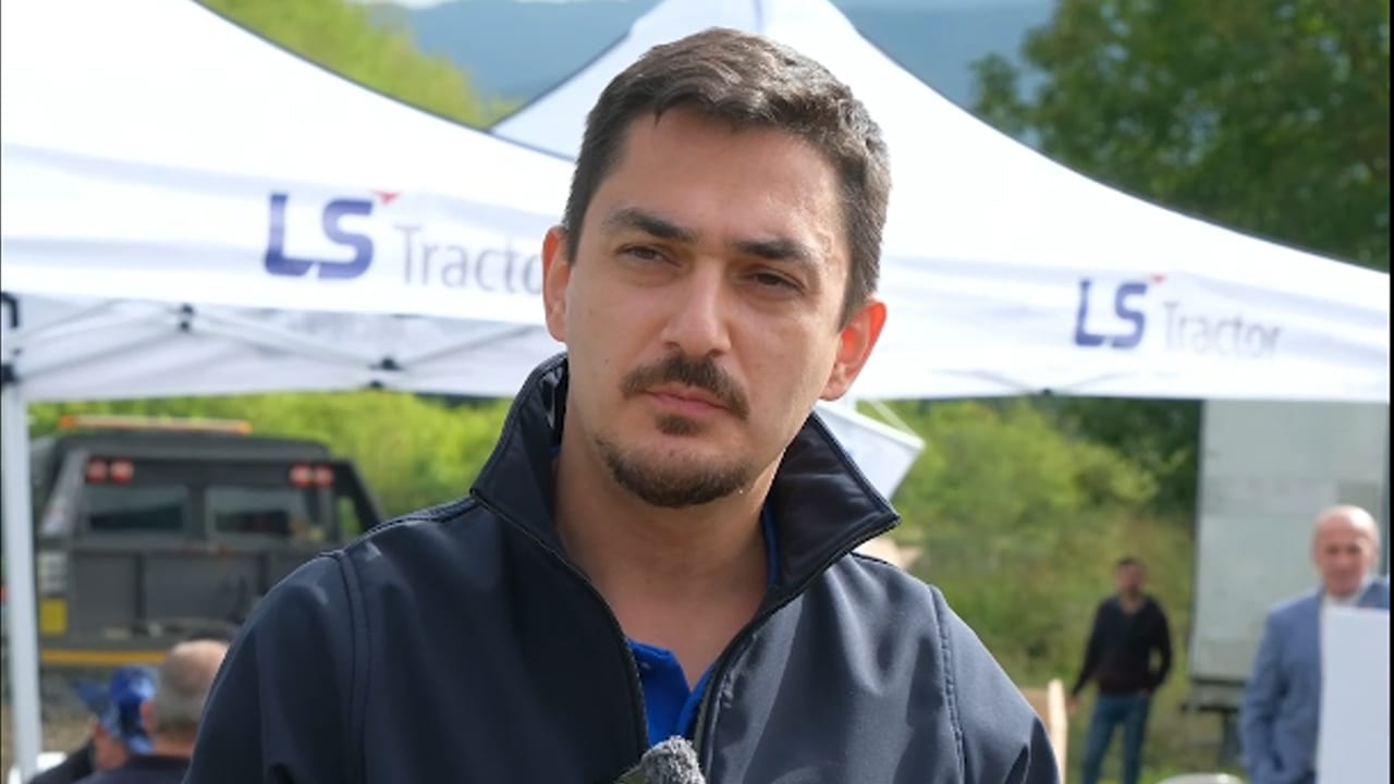 A man with a mustache and goatee wears a dark jacket in front of an "LS Tractor" tent.