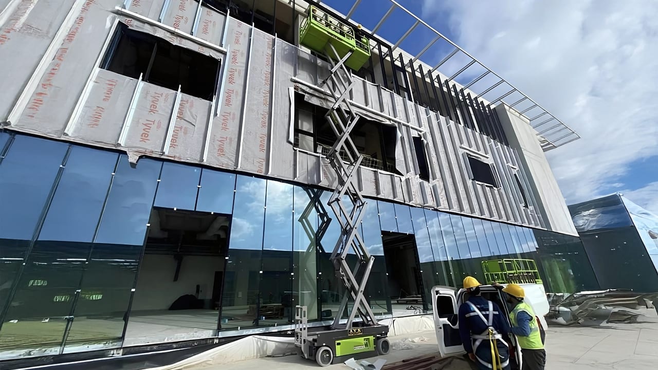 Building under construction with glass and Tyvek facade, scissor lift, and workers.