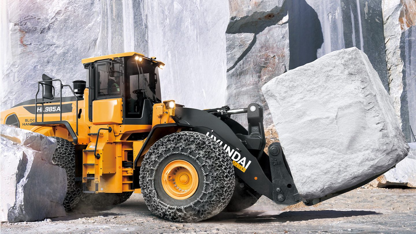 Yellow Hyundai wheel loader with chain tires carrying a large rock in a quarry.
