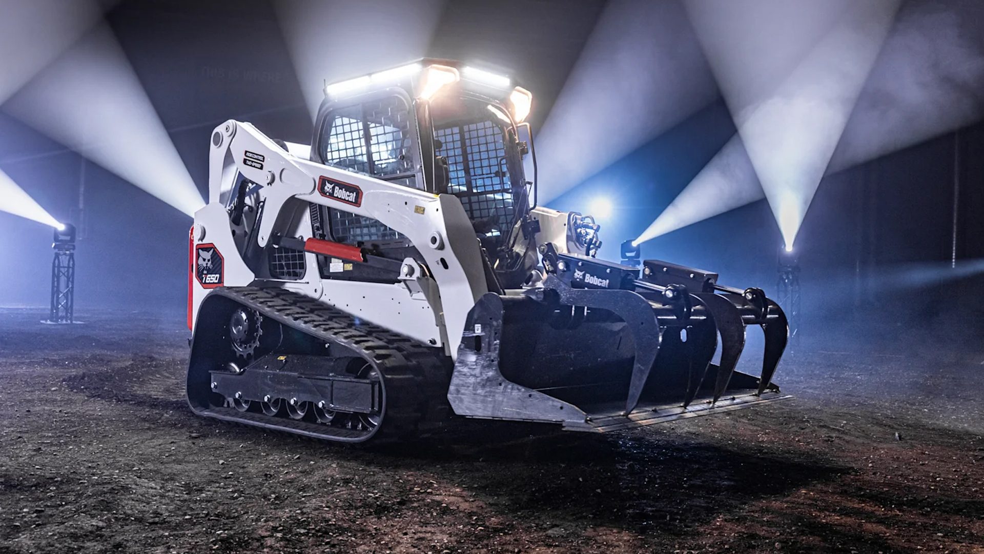 A white Bobcat compact track loader with a grapple attachment, illuminated by spotlights in a dark setting.