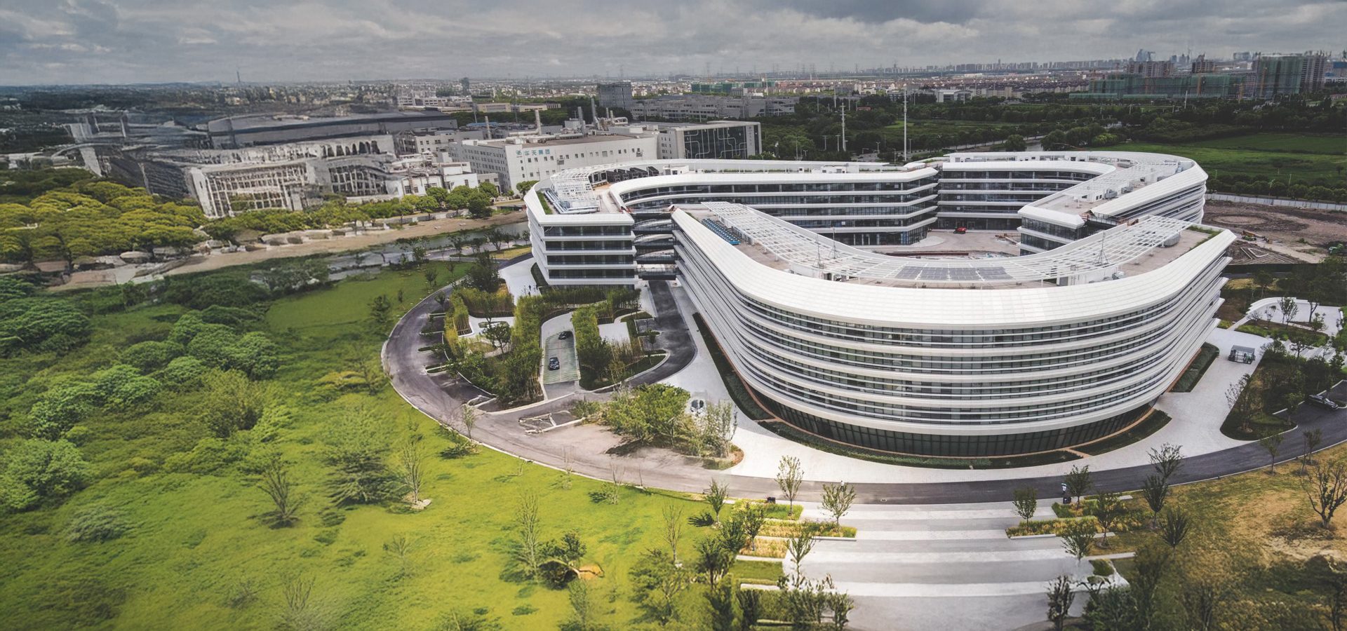 Aerial view: Modern, curved white building complex amid green landscape, city in background.