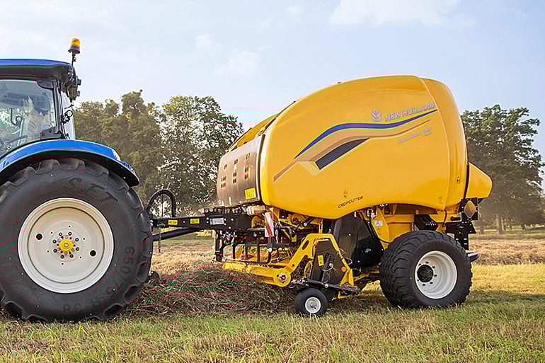 A blue tractor pulls a yellow New Holland round baler across a field, harvesting crops under a clear sky.